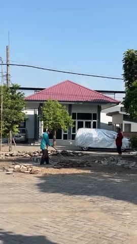 A person in a turquoise shirt and black pants is using a rake to clear debris from a paved area in front of a building. The building has a red roof and white walls. A car covered in a silver tarp is parked to the right of the building. Another person in a red shirt is standing to the right of the covered car. Trees are visible on the left and right sides of the scene. The sky is clear and blue.