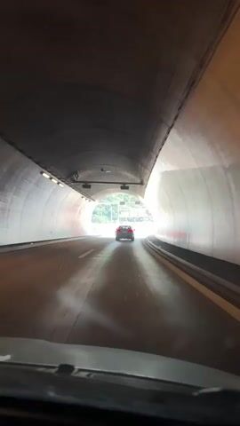 The video shows a first-person view from inside a vehicle driving through a tunnel and then emerging onto a highway. Inside the tunnel, the walls are illuminated by overhead lights. A red car is visible ahead. As the vehicle exits the tunnel, the scene transitions to a sunny day with a clear blue sky and some clouds. The highway is surrounded by green, rolling hills and mountains. There are other vehicles on the road, including the red car that was seen in the tunnel. Overhead signs and barriers line the highway.