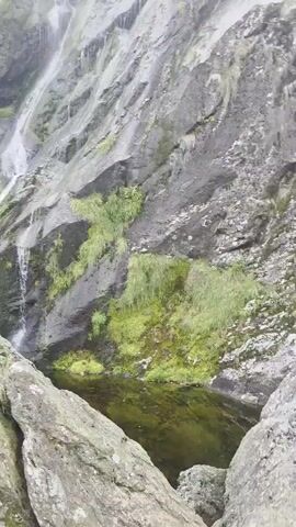 A wide shot of a tall waterfall cascading down a moss-covered rocky incline is captured. At the base of the waterfall, a small pool of water is visible. In the foreground, a person stands on a boulder, looking up at the waterfall. The person is wearing a dark t-shirt with a yellow stripe on the sleeve and dark pants. The environment is lush and green, with trees and vegetation visible on the upper slopes of the hill. The lighting is overcast, suggesting a cloudy day.