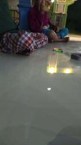 The video captures a dimly lit outdoor scene at night. The foreground is dominated by a smooth, light-colored tiled floor that reflects the ambient light. In the mid-ground, two individuals are seated on the floor. One person, wearing a maroon shirt and blue checkered shorts, is leaning against a blue pillar and appears to be smoking. This individual is facing away from the camera. To their left, another person is lying down, covered by a patterned cloth in red, white, and blue. In the background, a structure with green and white ornate trim is visible, possibly part of a building or a decorative archway, illuminated by yellow lights. A bicycle is parked near this structure. A small metal ashtray and a lighter are on the floor between the two seated individuals. The overall atmosphere is relaxed and informal.