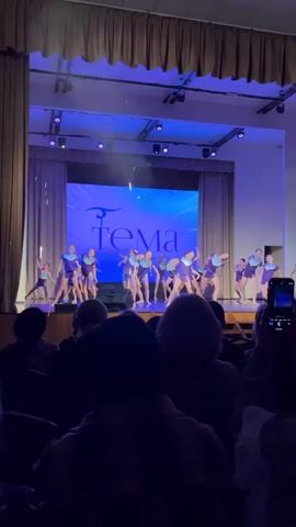 From a low-angle viewpoint, a performance is underway on a brightly lit stage. A group of young girls, dressed in purple leotards, dance in synchrony, striking dynamic poses and moving together. Their performance is against a backdrop featuring a large screen displaying the word "Tema" in Cyrillic script, accompanied by a stylized graphic. The stage is draped with heavy curtains, and illuminated by blue stage lights that add to the visual ambiance. The audience watches the show, their silhouettes visible in the foreground as they are mostly obscured by the darkness. Some audience members are recording the performance on their phones.
