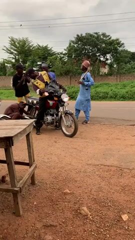 A group of men and motorcycles are situated on a dirt patch adjacent to a paved road under an overcast sky. In the center, a man wearing a yellow reflective vest over a dark shirt and a black cap is seated on a red motorcycle. To his left, a man in a black shirt and yellow pants holds a smartphone, apparently recording. Further left, partially obscured by a wooden table in the foreground, another man in a brown outfit is crouched or lying next to a white motorcycle. Behind the red motorcycle, a man in a blue shirt and black cap stands. On the far right, a man in a blue traditional outfit and a red cap stands, holding a long stick-like object. In the background, another motorcycle with a rider is visible on the paved road, moving away. Trees and a low wall are visible behind the road. An engine sound is audible.