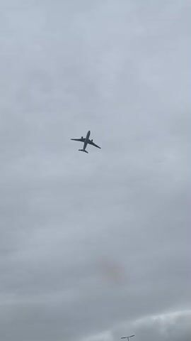 A wide shot captures an airplane flying against a cloudy, overcast sky. The plane is seen from below, its silhouette dark against the gray clouds. A small utility pole is visible in the lower part of the frame, suggesting the viewpoint is at ground level. The airplane appears to be in flight, steadily moving away from the viewer.