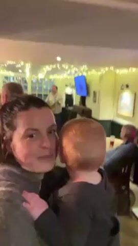 A woman is holding a baby in a pub.  The baby has red hair.  A man is playing an acoustic guitar in the background.  Several other people are in the pub as well.  String lights are strung around the ceiling.