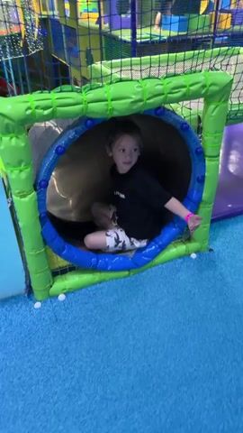 The video shows a young child going down a tube slide in an indoor playground. The child is wearing a black t-shirt and patterned shorts. They have a pink wristband on their left arm. The playground appears to be colorful, with blue and green padded structures surrounding the slide. The slide itself is a shiny, metallic tube. The child's face is visible in some shots, showing a mix of excitement and a slight grimace as they slide. The overall setting is an indoor play area, likely a children's entertainment center, judging by the colorful and soft structures. The lighting within the tube is somewhat dim, with brighter light visible at the end of the slide, suggesting it leads to an open area. There is no visible text in the video. The activity is a child's playful descent down a slide.
