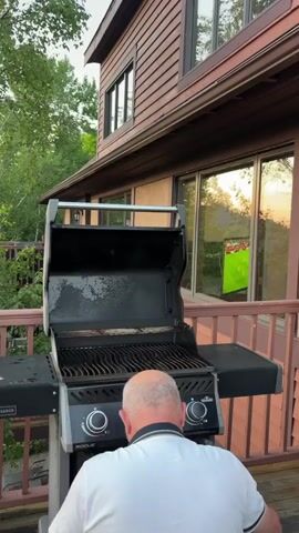 The video begins with a close-up of a man standing in front of a barbecue grill on a wooden deck. The grill is open, and the man is looking down at it. The camera then pans to the left, revealing a lush green forest and a sky with streaks of orange and pink, indicating either sunrise or sunset. The scene transitions to a wider shot of a picturesque town nestled beside a large body of water, surrounded by rolling hills. The sky is filled with dramatic clouds, and the water reflects the warm hues of the sky. The video ends with a slightly closer view of the town and the sunset.