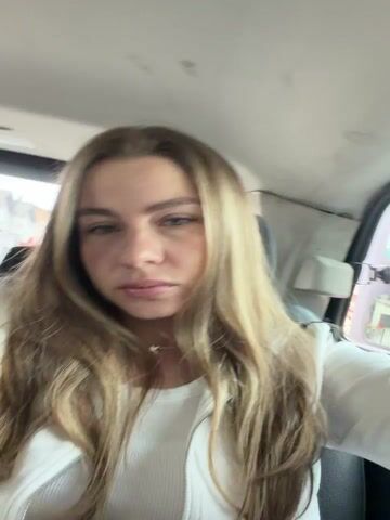 The video shows a young woman with long blonde hair sitting in the passenger seat of a vehicle. She is wearing a white hooded jacket over a white tank top and dark leggings. The woman is looking at the camera and occasionally touching her hair or face. The camera appears to be mounted on the dashboard or a similar position, looking towards the passenger side. The interior of the vehicle is visible, including a black seat and part of the window. Outside the window, trees and a building are visible, suggesting an outdoor setting. The lighting is natural and appears to be daytime. The video is filmed from a first-person perspective, with the woman occasionally turning to look out the window or adjusting her position. The overall mood is casual and relaxed, with the woman appearing to be on a journey or enjoying a drive.