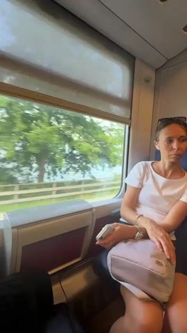 A woman sits on a train, looking out the window. She is wearing a white t-shirt and dark shorts. She holds a phone in her left hand and has a light-colored purse on her lap. The train is moving, and trees and water can be seen passing by outside the window. The window is covered by a roller blind.