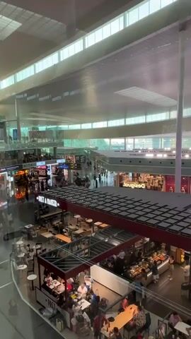 A wide-angle, high-angle shot looks down into a busy airport terminal. In the foreground, several tables with people seated around them are visible. Some are eating and drinking, while others appear to be conversing. A food stall with a display of food items is set up, and staff members are visible behind the counter. Above this area, a dark, slatted roof structure extends across the frame.

In the mid-ground, a large, illuminated digital billboard hangs, displaying an advertisement for "Shopping" with an image of a sandwich. Beyond this, another level of the terminal is visible, featuring shops with illuminated signs, including "Desigual." People are seen walking through this area, and more seating and dining areas are present.

In the background, the vast ceiling of the airport terminal is visible, with skylights allowing natural light to filter in. The overall environment appears modern and well-lit, indicative of a contemporary airport facility. The scene is populated with numerous individuals, suggesting a time of peak activity.