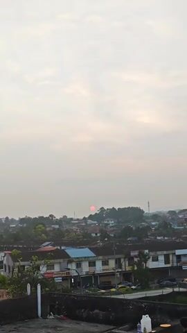 A time-lapse video shows a hazy sky with the sun setting behind a residential area. The sun appears as a small, pinkish-red orb. The residential area consists of rows of houses with red, blue, and white roofs, interspersed with green trees. In the foreground, there is a concrete structure with some debris and a white plastic container. The video captures the gradual descent of the sun as it approaches the horizon, with the sky becoming slightly darker and the overall scene fading.