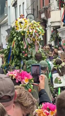 A crowd of people are gathered in a street to observe a parade. Two large, leafy figures covered in greenery and flowers are the main focus. The figures have floral crowns on top. People in the crowd are holding up their phones to record the event. One person in the crowd wears a green beret and has green paint on their face. Some in the crowd hold bouquets of flowers.  The street is lined with buildings. A clock on one of the buildings reads 3:16.  Colorful ribbons and banners are visible in the background. The overall atmosphere appears festive and celebratory.