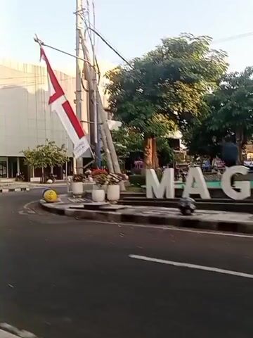 The video shows a street scene with a large, modern building in the background. Indonesian flags are visible, attached to poles and fluttering in the breeze. There are trees and planters along the sidewalk, and streetlights with ornate lamps. A traffic light is visible on the left side of the frame. The road is paved, with white lane markings. The sky is clear and bright, suggesting it is daytime. There are a few white spherical bollards on the sidewalk, and a red cylindrical postbox. Some people are visible walking on the sidewalk in the distance. The camera is moving, giving a sense of a slow pan or a drive-by.