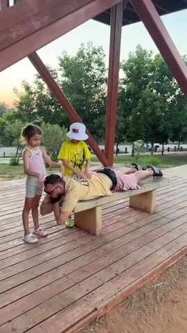 A man is lying face down on a concrete bench outdoors. Two children are standing over him. A young girl on the left is touching the man's head with both hands. A boy wearing a yellow shirt and a white hat is standing to the right of the girl and is also touching the man's head and shoulders with both hands. The man is wearing a tan t-shirt and pink shorts. The girl is wearing a pink and white patterned tank top and grey shorts. The boy is wearing a yellow t-shirt and white hat. The bench is on a wooden platform. In the background, there are trees and a cloudy sky at dusk.