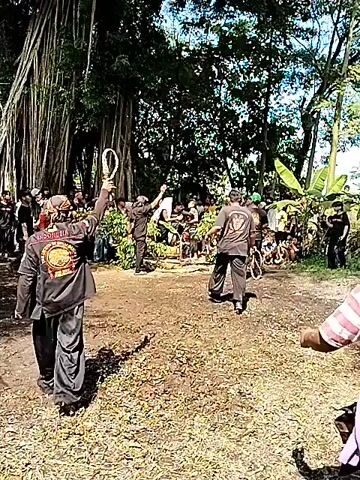 The video depicts a group of people gathered in what appears to be a park or forest area. There are tall trees and lush greenery visible. Several individuals are dressed in black shirts with logos on the back, others wear casual clothes like striped shirts and sarongs. One person is holding a rope or looped object above their head. The people are walking or standing, interacting with each other, seemingly part of an event or gathering. The overall atmosphere seems communal and festive.