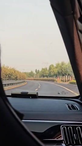 The video shows a view from inside a car, looking forward through the windshield. The road ahead is a winding highway with trees lining both sides. The sky is bright and hazy, suggesting daytime. A dashcam is visible on the dashboard, and part of the car's interior, including an air vent, is also in frame. The car appears to be in motion, as indicated by the road stretching out ahead. There are no other vehicles or people clearly visible in the immediate vicinity.