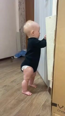 The video shows a baby standing while holding onto a wall for support. The baby is wearing a black long-sleeved bodysuit and a diaper. The baby is barefoot on a light brown wooden floor. The wall is white, and next to it is a cardboard box with the word "СТУЛ" printed on it. In the background, there is a radiator, a wall with a decorative design, and a blue stuffed animal lying on the floor. The baby looks at the camera and smiles.