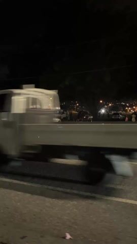 At night, a silver pickup truck is parked on the side of a road near a grassy area. Other cars are parked nearby, and a few people are milling around. The cityscape is visible in the distance, illuminated by many lights.