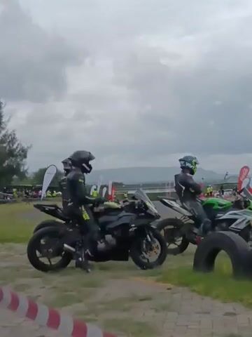 The video shows a group of motorcycle racers at a racetrack, waiting for a race to begin. The racers are dressed in black racing suits and helmets, and they are sitting on their motorcycles. The motorcycles are lined up along the edge of the track, and there are tires along the side. In the background, there are tents, flags, and people standing around the track. The sky is cloudy and overcast.