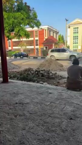 Here is a description of the video:
The scene is captured outdoors on a bright day with clear skies. A man, dressed in muted tones, sits on the ground, his back facing the camera. He is positioned to the right of a mound of dirt and bricks. The foreground is filled with a rough, speckled surface, possibly concrete. To the left, a red structural element is visible, likely part of a building or shelter from which the shot is taken.

In the background, across the street, stand a few buildings. The architecture is modern with clean lines, and they are painted in light colors. There are some trees with vibrant green leaves. A vehicle appears to drive past in the distance.