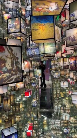 The video opens with a shot of an immersive art installation that appears to be a room lined with mirrors, creating an infinite reflection effect. Suspended from the ceiling are numerous illuminated cubes, each displaying a different famous painting. The art ranges from classic European masterpieces to more modern pieces. In the center of the room, a person stands on a reflective platform, holding a phone and appearing to take a picture or video of the surroundings. The camera slowly pans and zooms, revealing more of the art-filled environment, with reflections of the cubes and the person creating a dizzying and expansive visual experience. The lighting is soft and varied, with some cubes emitting a warm glow and others a cooler light, accentuating the artwork. The overall impression is one of a surreal and captivating art exhibition.