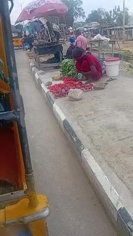 The video shows a street market scene taken from inside a yellow tuk-tuk. A woman in a red dress is crouched on the sidewalk sorting a pile of red onions, next to a bunch of green leafy vegetables and some yellow produce. Behind her, vendors are selling goods under umbrellas. Other pedestrians are walking along the sidewalk, including two women carrying bags and bundles on their heads. One woman is carrying a baby or small child on her back. Various goods, including what appears to be dried fish, are displayed on tables. There are also several bags of yams on the sidewalk.