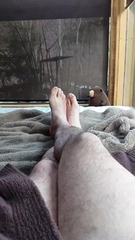 The video shows a person lying on a bed with their legs outstretched towards the camera. The legs are lightly hairy. The person's bare feet are visible, toes pointing upwards. The bed has a thick, textured gray blanket. A brown knit sweater lies to the left of the legs. Behind the bed is a window with a dark screen. Outside the window, trees are visible with bare branches. A brown stuffed beaver toy sits on the window sill. A loud noise is heard in the audio.