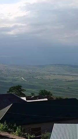 A vertical video displays a panoramic view of a rural landscape under a cloudy sky. In the foreground, the dark, sloped roofs of two houses are visible, one with a green-tiled surface and the other with corrugated metal. Partially obscured by the roofs are the tops of trees and a sliver of a building. Beyond the houses, the landscape stretches out, characterized by rolling green hills and fields. A winding road can be seen cutting through the valley. The sky is filled with large, grey clouds, with patches of lighter sky suggesting sunlight breaking through. The overall impression is one of a serene, expansive rural setting.