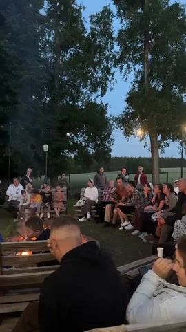 A group of people, including children and adults, are gathered around a campfire outdoors at dusk. They are seated on wooden benches and pallets, facing the fire. The scene is illuminated by the flames of the campfire and some ambient light from a string of lights hanging in the trees. The background consists of trees and a field. Some individuals are looking at the fire, while others are engaged in conversation.