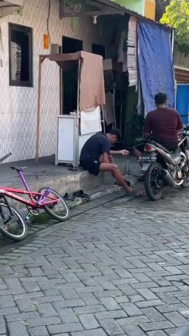 The video shows a young man sitting on a curb, hunched over with his head down, holding a cigarette and appearing to be smoking. He is wearing a black t-shirt and black shorts. To his right, a person sits on a black motorcycle facing away from the camera. The motorcycle appears to be parked on the street. In the background, there is a building with a tiled facade and what looks like a stall or small shop. There are also some objects leaning against the building, including what might be a ladder. The ground is paved with interlocking bricks. The overall environment appears to be an outdoor urban setting, possibly a street or alley.
