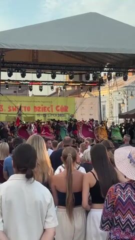 A wide shot captures an outdoor festival with a large stage and screen setup. A crowd of people watches a performance on stage, where individuals in vibrant, traditional costumes, possibly from Mexico, are dancing and playing instruments. A large screen displays a live feed of the performance. The atmosphere appears festive, with many attendees holding up phones to record the event. The sun is setting, casting a warm light on the scene.