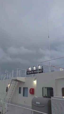 The video shows a close-up view of the side of a white ship. Above, a dark, cloudy sky suggests it might be evening or an approaching storm. A sign that reads "TETHYS HIGHWAY" is mounted on the ship's superstructure. Four lights illuminate the sign from above. To the left of the sign, a metal staircase with railings leads up. Several windows are visible on the ship's white exterior, one with a red fire extinguisher mounted beside it. A tall, thin antenna extends vertically from the ship's roof. In the foreground, a gray air conditioning unit or similar piece of equipment is partially visible. The camera is positioned at a low angle, looking upwards.