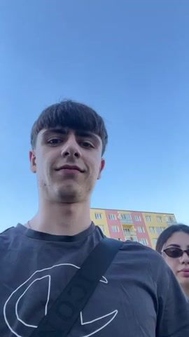 This is a short, selfie-style video featuring two young people walking outdoors on a sunny day. The primary subject is a young man in the foreground, wearing a dark grey t-shirt with a white Champion logo. He has short, dark hair styled in a bowl cut and is smiling broadly at the camera. He is also wearing a black strap across his chest.

Behind him, a young woman is visible, wearing a black sleeveless top and sunglasses. She has long, dark hair pulled back into a ponytail and appears to be talking or reacting to something off-camera.

The setting appears to be a residential area. In the background, a tall, multi-story apartment building with colorful facade is visible. Trees and a street with parked cars can also be seen. The sky is a clear, bright blue, indicating it is daytime, likely late afternoon or early evening given the lighting. The overall mood seems lighthearted and casual, with both individuals appearing relaxed and engaged.

No discernible text is visible in the video except for the Champion logo on the man's shirt.