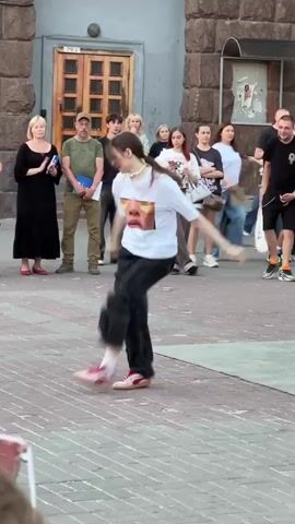 A young woman with long brown hair in a ponytail, wearing a white t-shirt with a graphic print, and baggy black cargo pants, is performing a dynamic dance routine on a paved outdoor area. She is in mid-movement, with her left leg extended forward and her right leg bent behind her. Her arms are in motion, contributing to the energetic performance. She is wearing pink sneakers with white socks.

Behind her, a group of people are observing the performance. To her left, a woman in a black dress stands with her hands clasped. To her right, a man in a black t-shirt and black shorts watches her dance. Other spectators, including young women and men, are visible in the background, some of whom appear to be holding phones to record the event.

The environment appears to be an urban public space, possibly a plaza or square, with a large building featuring a wooden door and stone facade in the background. The ground is paved with brick-like stones. The overall lighting suggests it is daytime, possibly late afternoon.