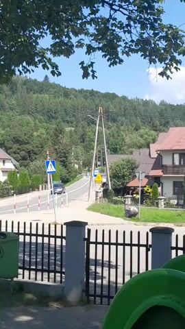 The video opens with a street view in a village or town. A black metal fence is in the foreground with green objects that appear to be playground equipment peeking out from behind it. Beyond the fence, a paved road curves to the left. A white crosswalk sign with a blue background and a white pedestrian symbol is visible on the left side of the road. A few cars are driving on the road, and there is a traffic light in the distance. On the right side of the road, there are houses with red tile roofs. Trees and hills covered in vegetation are visible in the background. The sky is blue and appears to be a clear day.