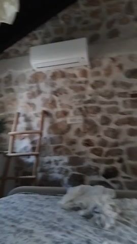 The video pans across a room with a stone wall. An air conditioning unit is mounted high on the wall, to the left of the frame. Below it, on the wall, is a light switch. To the right, there is a window with green shutters. A ladder is leaning against the wall to the left of the air conditioner. A crumpled piece of white fabric lies on the floor in the foreground. The ceiling is dark.