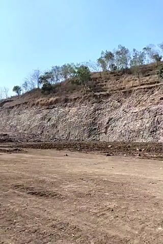 A drone's-eye view captures a natural landscape under a clear blue sky. In the foreground, a dusty, barren expanse of earth stretches out, marked by tire tracks suggesting vehicle passage. Behind this, a large, sheer rock face rises dramatically, its stratified layers clearly visible. The rock face is mostly devoid of vegetation, save for a few sparse bushes clinging to its upper edge. Above the rock face, a gentle slope continues upward, densely covered with trees and shrubs, their foliage sparse and dry, indicating a warm, arid climate. The overall scene depicts a rugged, undeveloped terrain.