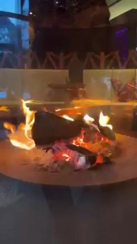 The video shows a close-up of a circular fire pit with burning logs and embers. The fire is active with visible flames. The pit is enclosed by glass walls. In the background, there is a seating area with cushions and a decorative wall feature. The lighting is dim, suggesting an evening or indoor setting. There is indistinct speech audible in the background.