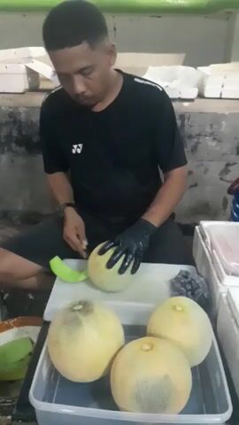A man wearing a black t-shirt and black gloves is sitting down and cutting a green melon. He is using a silver knife. There are three other melons in a plastic container in front of him. The background appears to be a market or food stall with white boxes and some hanging fabric.