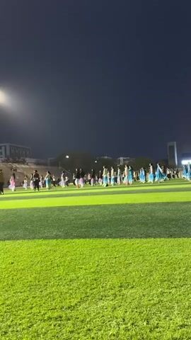 On a green athletic field at night, a large group of people are practicing dance movements. The field is lit by stadium lights, and tall buildings are visible in the background. The dancers are arranged in rows and columns, moving in a synchronized manner. Many are wearing light-colored tops, skirts or wraps in light blue, and sneakers, while others are wearing dark-colored clothing. A few individuals, seemingly instructors, are interspersed among the dancers. The camera captures a wide shot of the scene, focusing on the collective performance of the dance group.