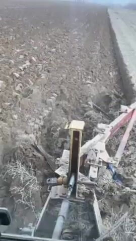 The video shows a view from inside a tractor as it plows a field. The plowing equipment is visible behind the tractor, churning up the soil. The field is expansive and the weather appears overcast. The tractor moves steadily, creating furrows in the earth.