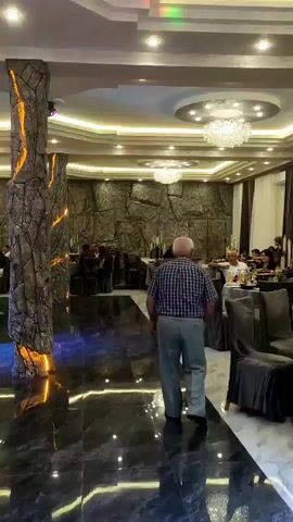 The video shows an indoor event, possibly a party or reception. The room is well-lit with chandeliers and ceiling lights. The walls are decorated with a stone-like texture in some areas and draped curtains in others. Tables are set with chairs, some of which are covered with fabric. There are several people present, including men, women, and children. In the foreground, a man in a checkered shirt walks away from the camera. To the right, a woman in a white shirt is visible, and further to the right, a child's head is partially seen. In the background, there are groups of people gathered around tables, some interacting with each other. There are also decorations such as flowers on a table. The floor in the foreground is dark and reflective. Green laser lights are visible on the back wall.