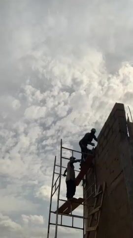 Two construction workers stand on a makeshift scaffolding system next to a partially built brick wall under a cloudy sky. One worker, wearing a green helmet, is laying bricks on top of the wall. The other worker stands below him on the scaffolding. The scaffolding appears to be a combination of metal and wooden structures.