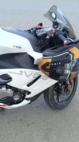 The video displays a white Voge motorcycle parked outdoors on an asphalt surface. The motorcycle is oriented with its right side facing the camera, and the shot is a close-up, focusing on the body of the motorcycle. The motorcycle features black and grey accents, as well as "VOGE" branding and "TECHNOLOGY" text. A gold and black helmet is visible resting on the seat of the motorcycle. The video pans slowly from the front of the motorcycle towards the rear, capturing details of the engine, exhaust system, wheels, and seats.