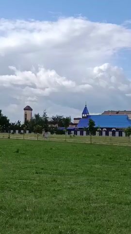 A vertical video displays a view of a building with a bright blue roof and white walls, adorned with dark trim. To the left of this building, a taller, slender tower with a brown roof is visible. In front of the structures, a black and white fence runs horizontally across the frame, with several small trees planted in a line behind it. The foreground is dominated by a large expanse of green grass. The sky above is blue with scattered white clouds. The lighting suggests daytime. There are no visible people in the immediate foreground.