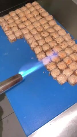 The video shows a close-up, overhead view of a blue cutting board where rows of small, square-shaped food items, possibly sushi rolls or appetizers, are arranged. A culinary torch with a bright blue flame is being used to sear the tops of these food items. The torch is held in the left side of the frame and is directed towards the food, creating a searing effect that lightly browns the surfaces. The food items appear to have a light-colored topping, possibly a sauce or cheese, which is caramelizing under the flame. The background is dark, with hints of metallic surfaces, suggesting a kitchen environment. The lighting is focused on the food and the torch, creating a dramatic effect. The action is continuous throughout the short clip.