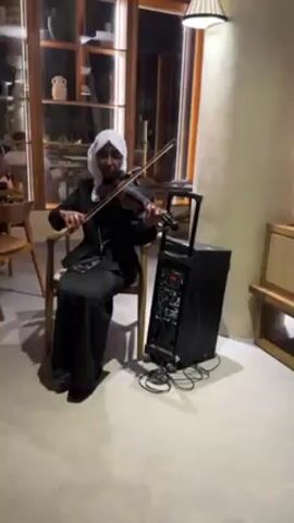 A woman seated on a chair is playing a black violin in what appears to be a restaurant. She is wearing a black suit, a white headscarf, and glasses. To her right is a black speaker on wheels with a control panel displaying some numbers. Behind her, a glass cabinet is visible, containing decorative items. The restaurant has light-colored walls and wooden furniture. Other people can be seen in the background. The room is illuminated by a hanging lamp.