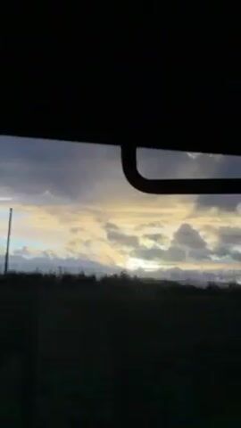 The video appears to be shot from inside a moving vehicle, looking out of the rear window and then the side window. Initially, the view out the back window captures a cloudy sky with hints of sunlight and a dark, indistinct horizon. The camera quickly shifts to the side window, providing a blurred view of the passing landscape, likely a field or grassy area. The overall lighting is subdued. The interior of the vehicle is visible through the window frame, appearing dark.
