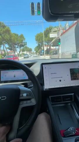 This media file provides a first-person perspective from inside a car, showing a view through the windshield and the car's dashboard. The driver's left hand is visible on the steering wheel. The dashboard features two screens: a digital instrument cluster on the left displaying vehicle speed and navigation information, and a larger central infotainment screen on the right. The infotainment screen shows a music player interface with the song title "Adrenalina" by Rels.

Outside the car, the scene depicts a road lined with trees and buildings, suggesting an urban or suburban environment. The sky is bright and clear, indicating a sunny day. Traffic is visible on the road ahead, including other cars. A traffic light is illuminated green. Visible text includes the song title "Adrenalina" on the infotainment screen, and a Greek sign that reads "ΧΑΖΑΠΟΝΟΧΕΥ" above a traffic light. The overall mood appears relaxed and ordinary, consistent with daily driving. The location is identified as Nea Kerassia, Greece.