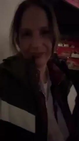 A woman with brown hair and a light complexion looks at the camera in a selfie video. She is wearing a grey jacket with a white stripe and a maroon collar. The background appears to be a dimly lit indoor space with people sitting and moving around, and some red accents in the lighting or decor. There seems to be loud music or a gathering occurring. The woman smiles slightly and seems to be enjoying the atmosphere.