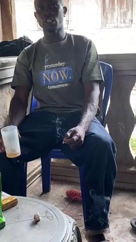 A person wearing a green t-shirt with the words "yesterday", "NOW.", and "tomorrow" printed on it, is sitting in a blue chair and smoking. The person is wearing dark blue jeans. The video is filmed from the chest up to the top of the head, showing the person's arms and hands holding a cigarette. Smoke is visible rising from the cigarette. The background is blurred but appears to be an indoor or covered outdoor setting with wooden structures visible.
